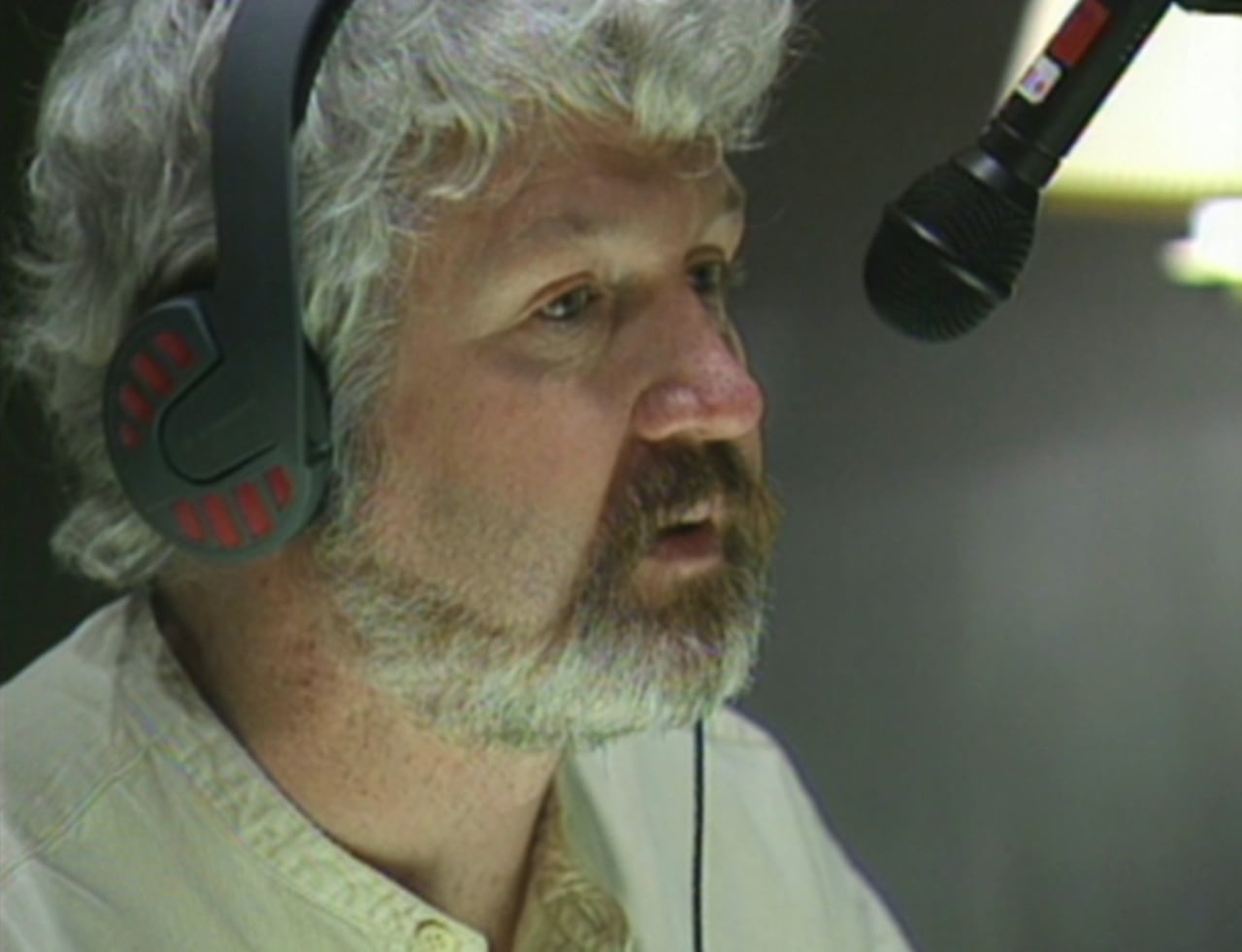 A man with a beard and curly white hair speaks into a microphone above his noise.