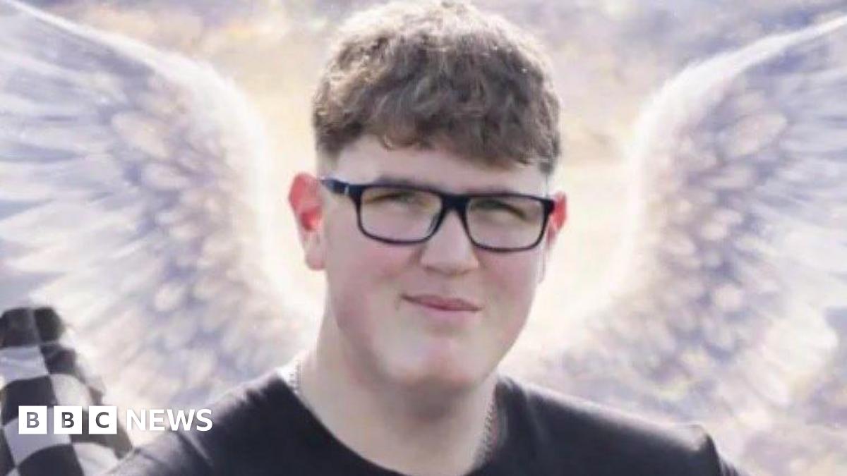 An image of a young man with glasses and short curly brown hair. He is looking at the camera and smiling. Behind him are some animated angel wings, a black and white chequered racing flag and a tyre.