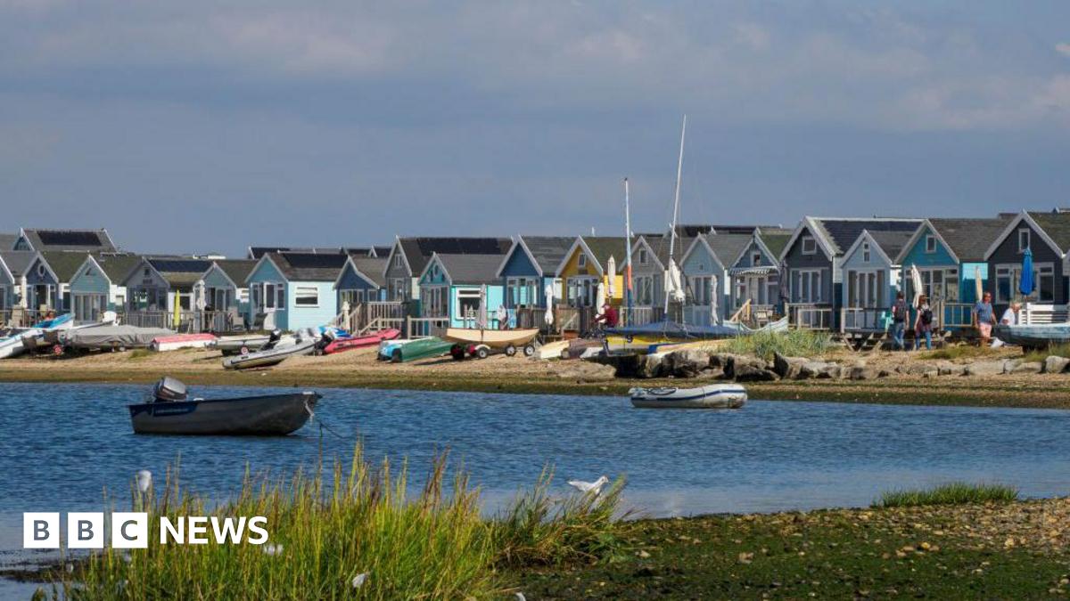 A row of brightly coloured beach huts stand on a sandy beach next to some small dunes. There are picnic benches outside the huts. The sky is blue and it is a sunny day. There is a body of water in front of the beach with some plant life on the near shore. There are several small boats in front of the huts and two in the water.