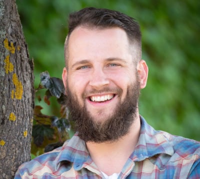 A portrait of a smiling man standing with his arms crossed. He's outdoors, leaning against a tree.