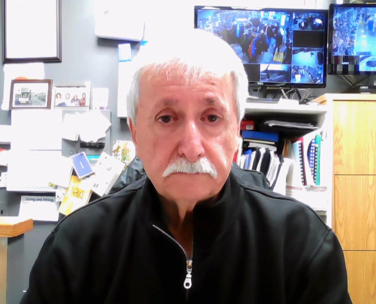 A man wearing a black quarter zip sweater sits in an office.