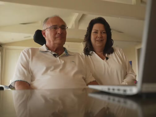 A man in a wheelchair looks at a laptop screen while a woman sits next to him. 