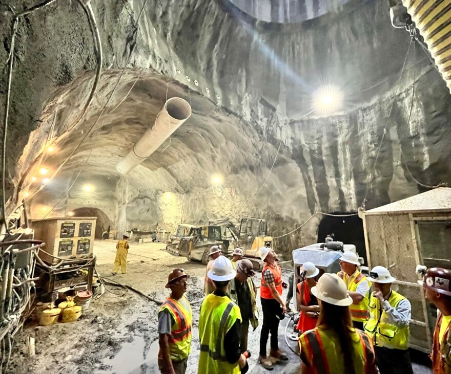 People in safety hats and vests stand underneath a chamber building underground. 