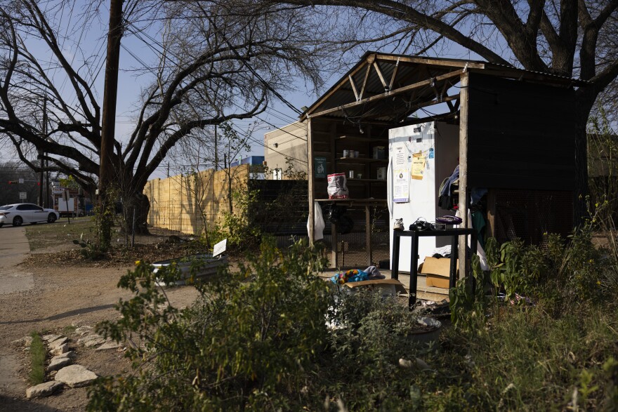 A refrigerator sits under a covered structure with bushes in the foreground beside a street.