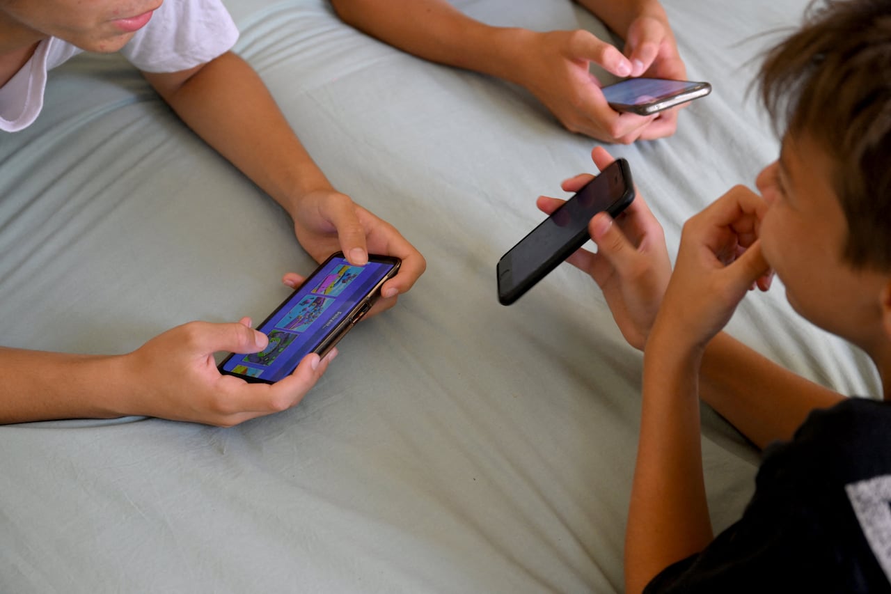 Close-up image focused on smartphones in the hands of three teens, one person at right turning their screen to show the two others sitting opposite.