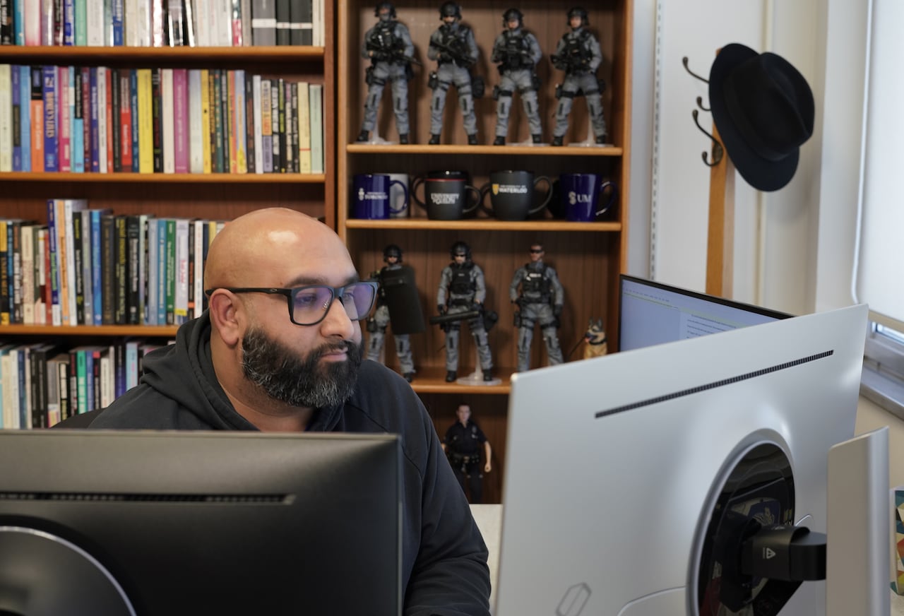 A man in glasses with a dark hoodie on sits at a desk with computers.