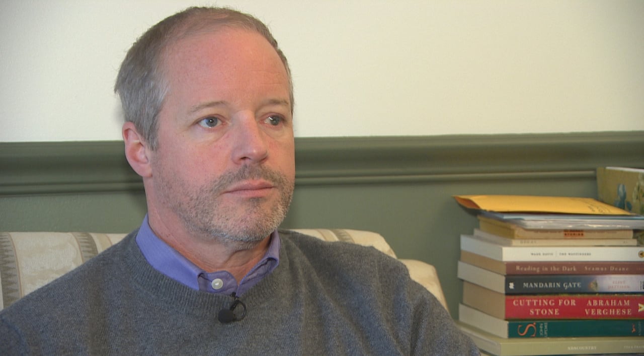 A man in a sweater sits with books stacked beside him.