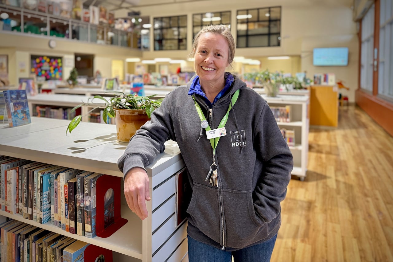 A woman with blonde hair in a grey hoodie leans against a bookshelf. She smiles.