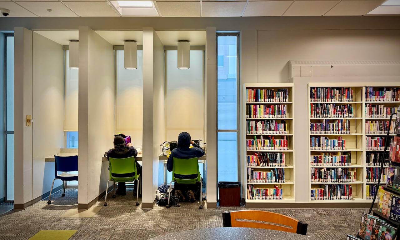 Two people sit at desks in the libraries with their belongings. Their backs are to the cameras and they are surrounded by bookshelves.