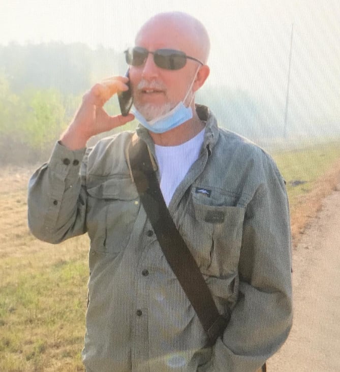 A man in sunglasses stands on a roadside with a phone pressed to his ear.