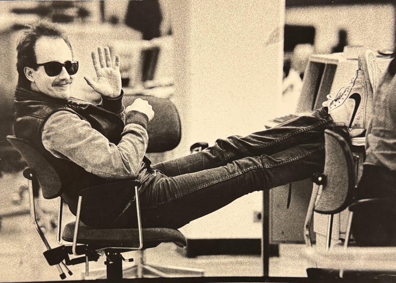 A black-and-white image of a young man in sunglasses with his feet up at a desk, waving at the camera.