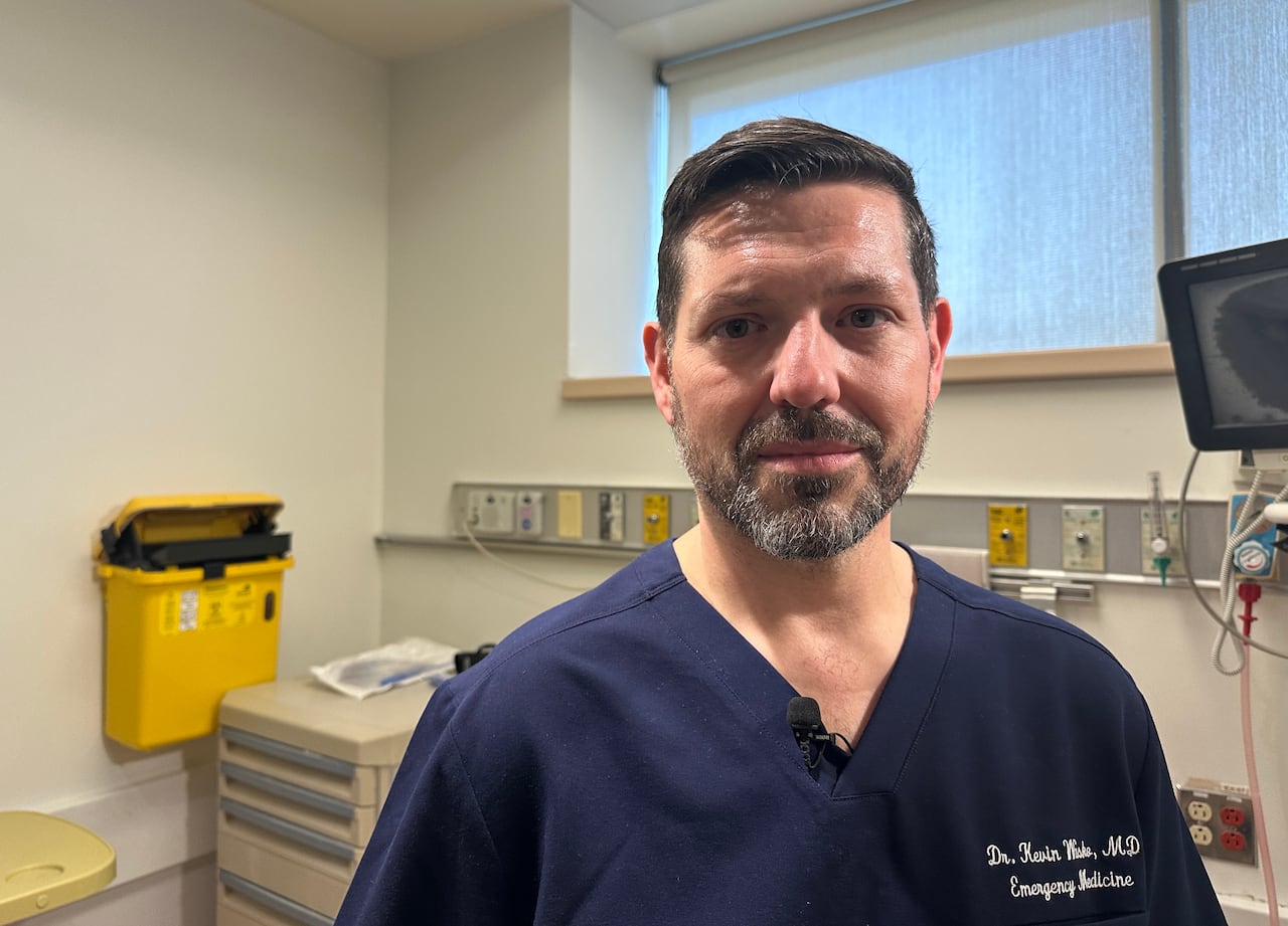 A man with brown hair and a grey beard wearing navy scrubs emblazoned with Dr. Kevin Wasco, MD, Emergency Medicine
