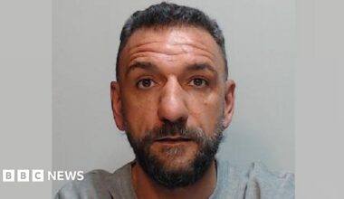 a police mugshot of a man with short black hair and a black beard. He is wearing a grey t-shirt