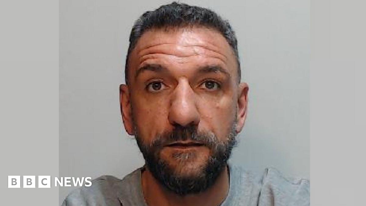 a police mugshot of a man with short black hair and a black beard. He is wearing a grey t-shirt