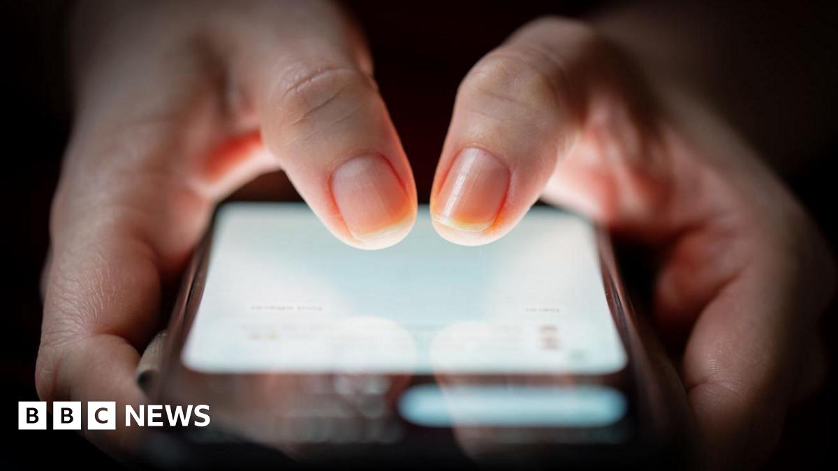 Two hands holding a smartphone. The screen of the phone is lit up and the background is dark. Both thumbs are poised to type in a number.