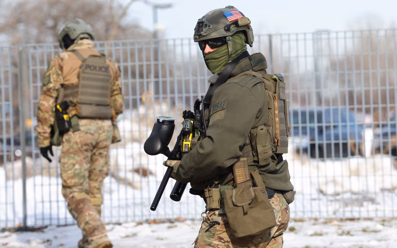 A masked federal agent holding a weapon is seen against a backdrop of a metal fence. 