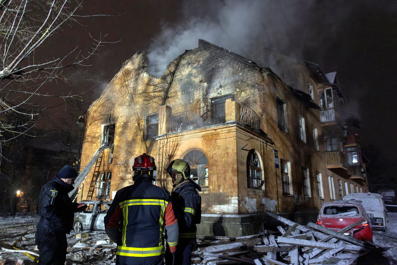 Firefights stand in front of a damaged brown brick building at night. Smoke rises from the building. 