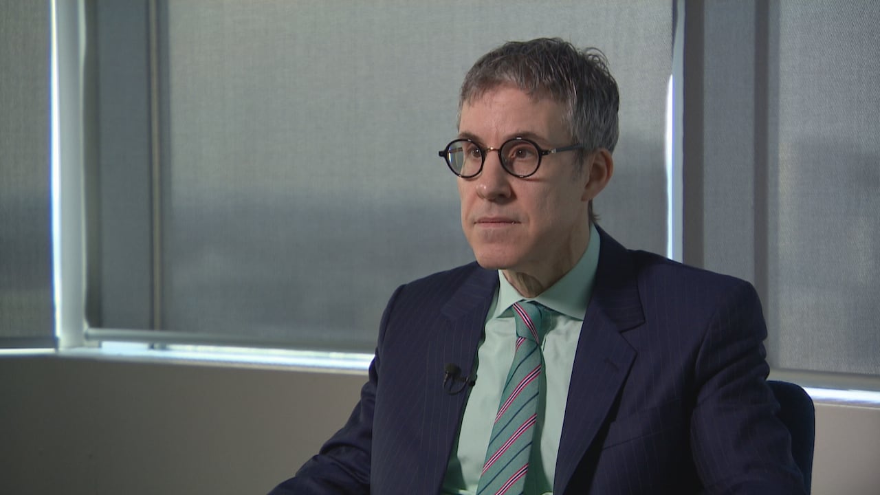 Man with grey hair and dark, round eyeglasses wears a dark blue suit and light blue dress shirt