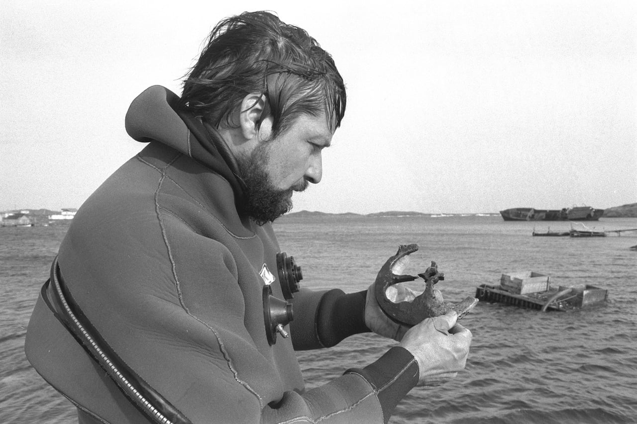 A black and white photo of a man next to the ocean holding a rounded object in his hands.