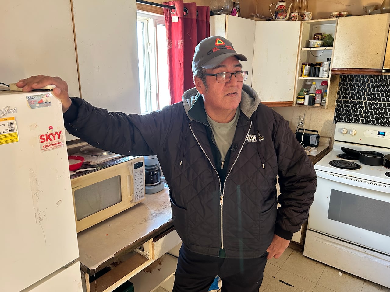 A man in a black jacket leans on a fridge as he looks out at his nephews' home.