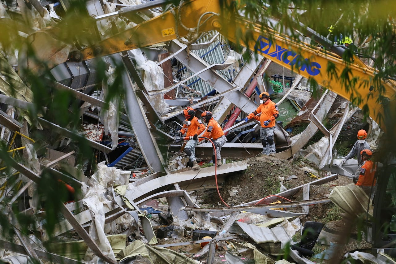 Rescuers in orange uniforms and protective gear and PPE search through rubble and garbage with a huge crane seen above them 
