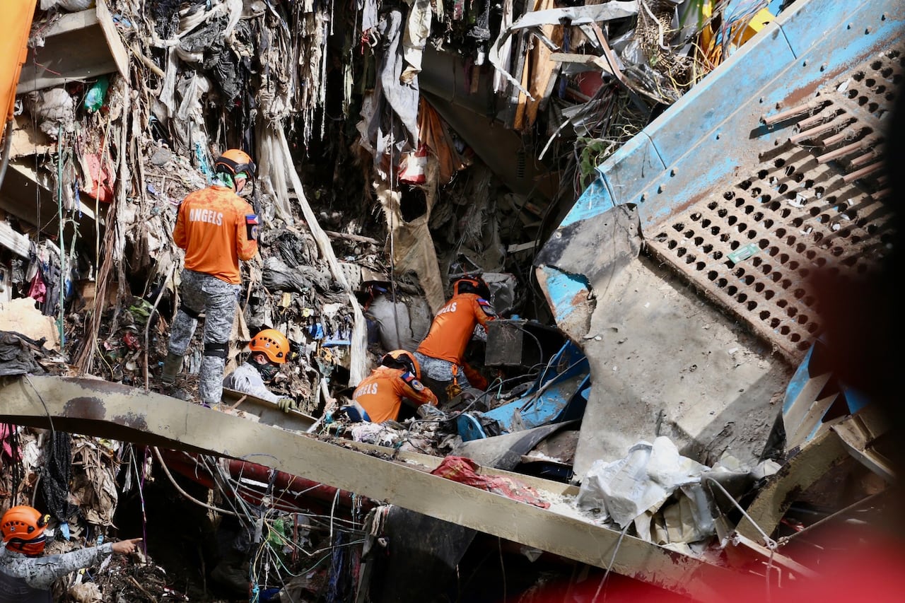 Rescuers in uniforms and protective gear, including helmets, search through heaps of garbage and destroyed infrastructure following a trash avalanche