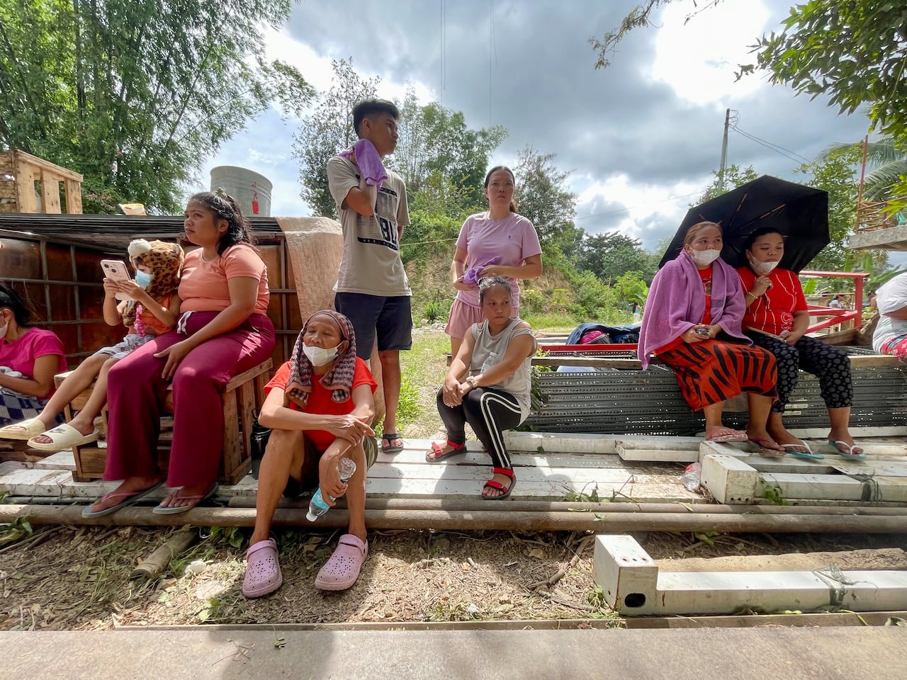 Group of people, ranging from children to elderly women, sit, waiting, with umbrellas and towels cooling them off.