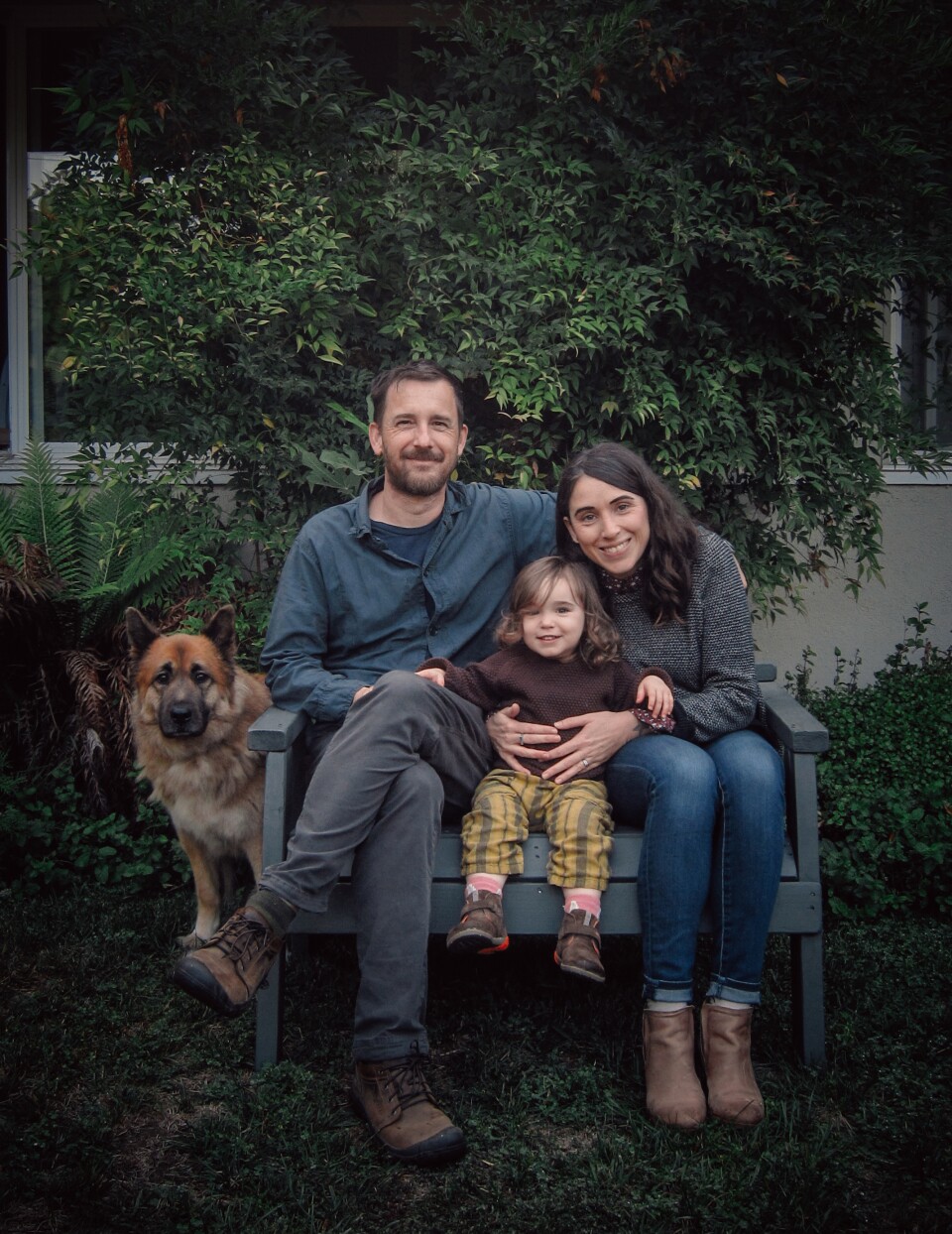 A family of three -- a man, a woman and child -- poses on a bench outside next to a brown large dog with pointed black ears. 