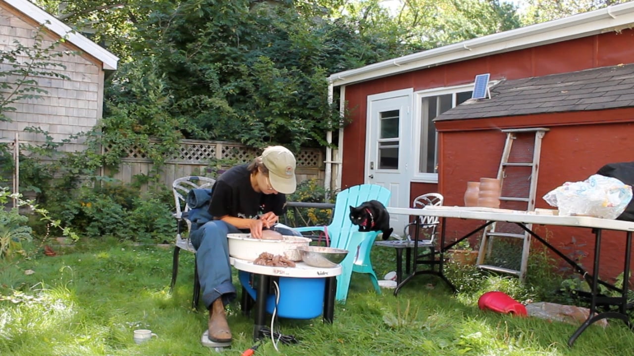 A person throws pottery on a wheel, while a little black cat watches