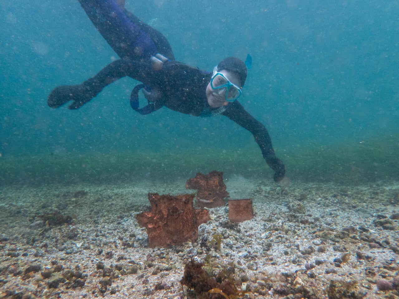 A person in snorkeling gear in the water. Below, pots made form clay lay on top of the ground. 