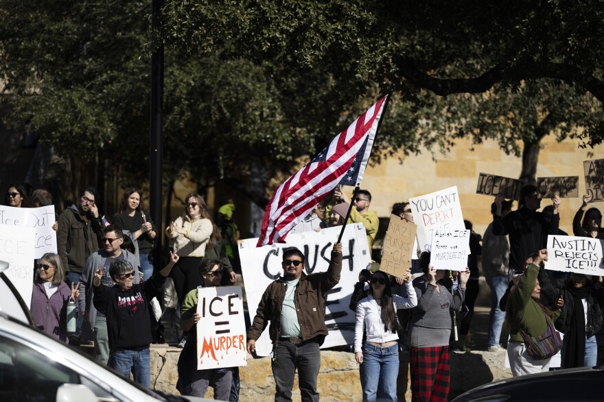 People gather holding signs against ICE  in front of city hall as cars go by. A man in the center waves a U.S. flag.