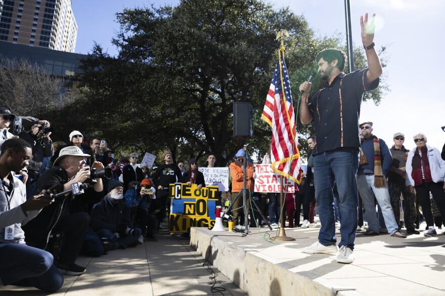 A man stands on a small podium and speaks to a crowd of media and protesters. A U.S. flag hangs nearby and some people in the crowd hold anti-ICE signs. 