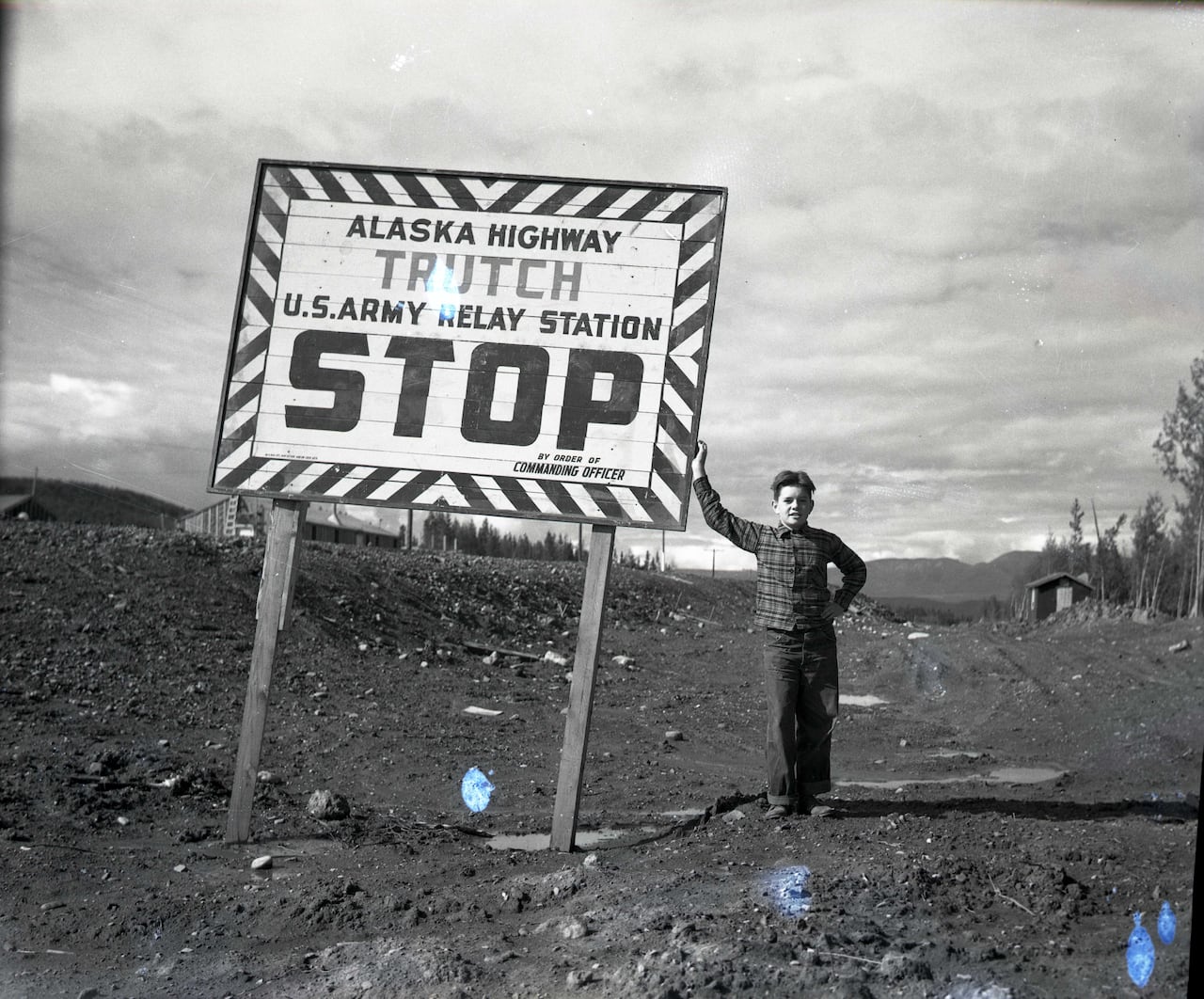 A boy stands next to an Alaska Highway relay station sign at Trutch, Mile 195.