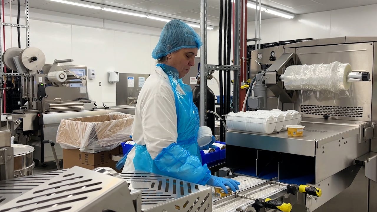 An employee wearing a hair net, lab coat, plastic apron and gloves hand packages Nuts For Cheese dips. 