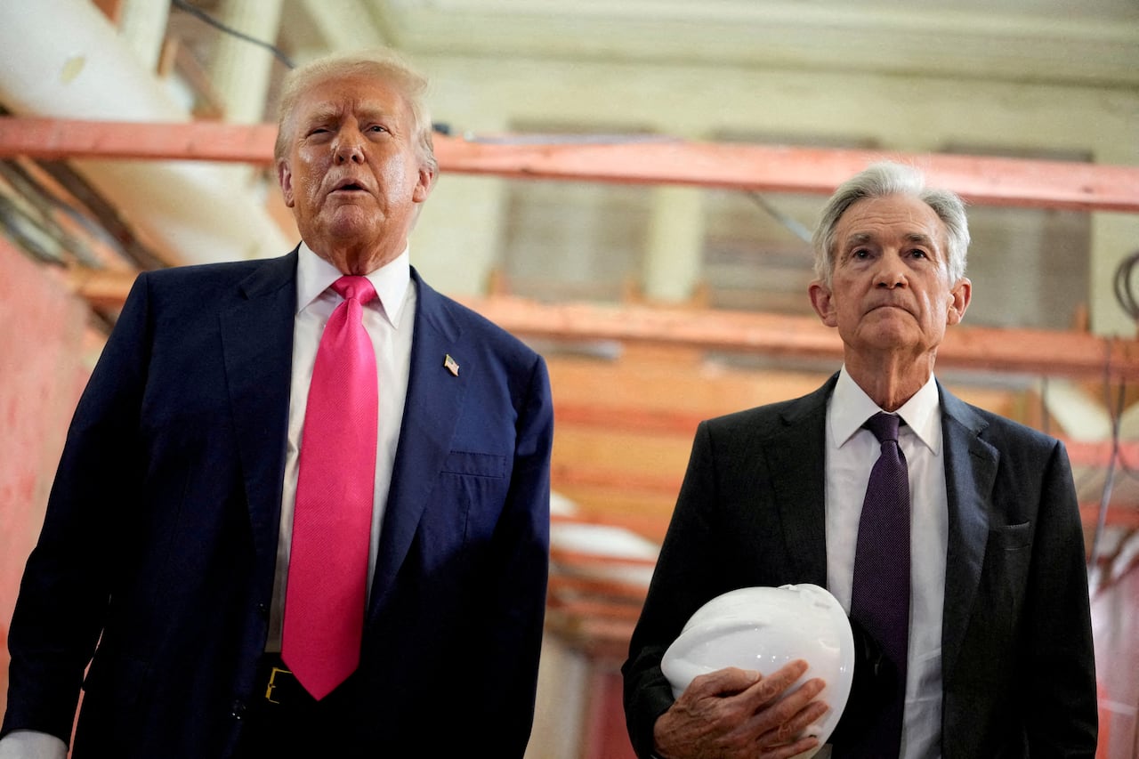 Two older men stand in a construction zone. The man on the right holds a hard hat.