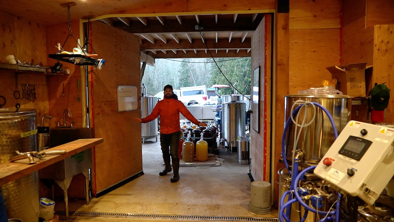 A man stands in the doorway of a building filled with steel drums and barrels for making cider. 
