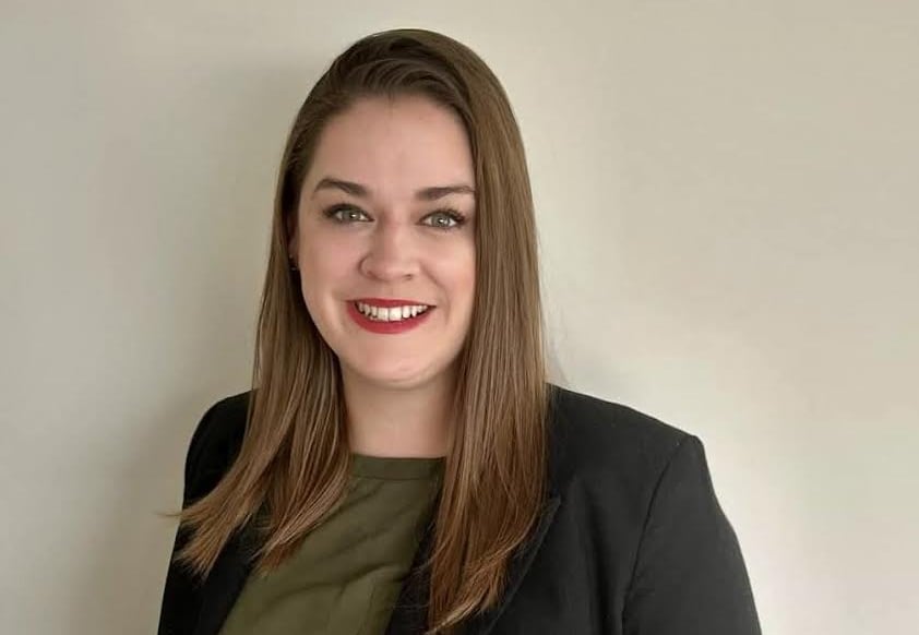 A woman with long brown hair and a black blazer smiles at the camera with a beige wall behind her. 