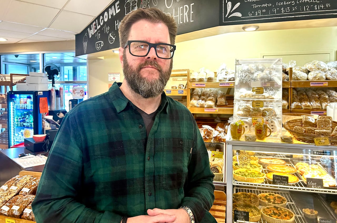 A man with brown hair and a beard smiles at the camera while standing in a bakery with food displayed behind him.