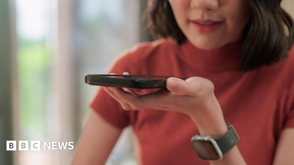 A woman wearing a red top speaks into the speaker of a smartphone she is holding in front of her.