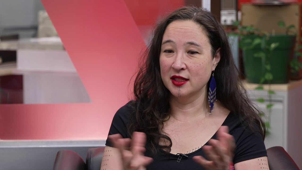 A woman with dark hair and facial tattoos speaks 