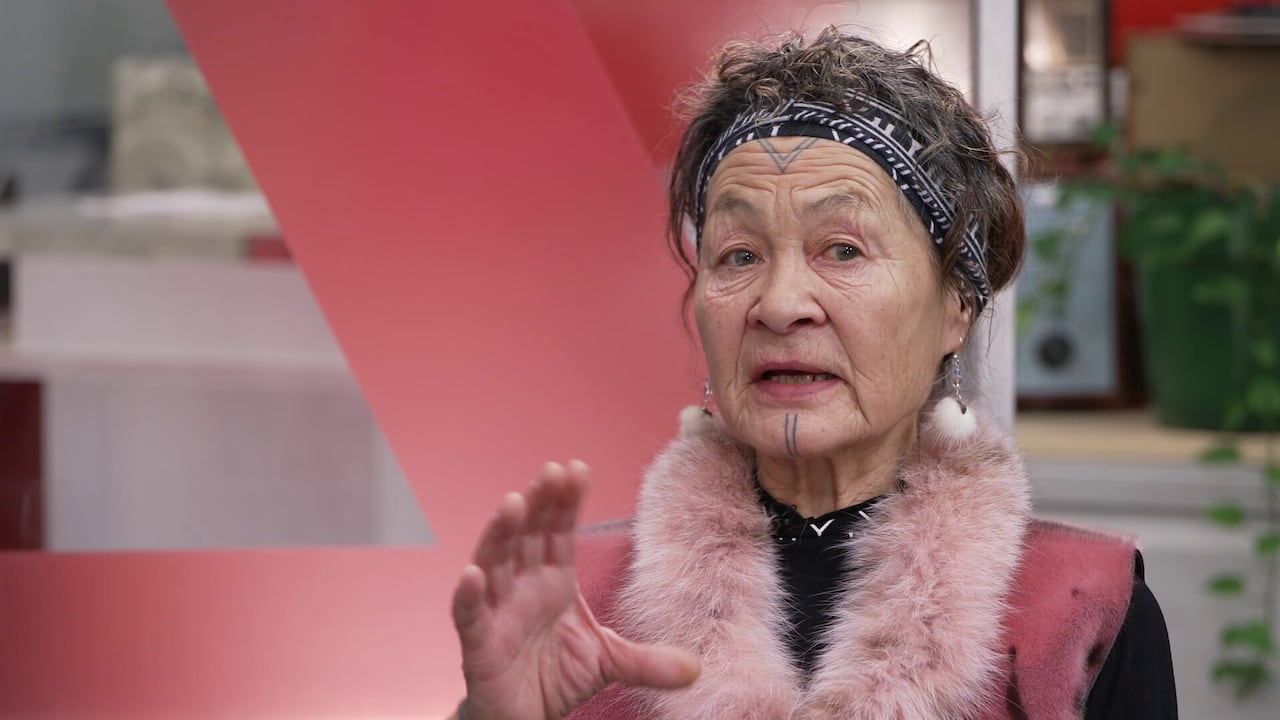 A woman with a chin tattoo and a fur vest speaks to the camera. 