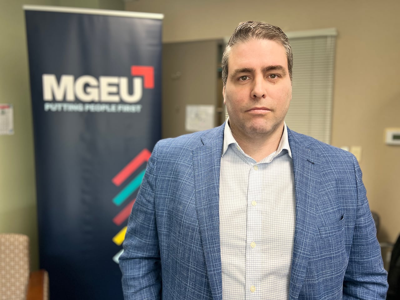 A man in a blue blazer stands in his office, as a banner with the "MGEU" logo is situated some distance behind him.