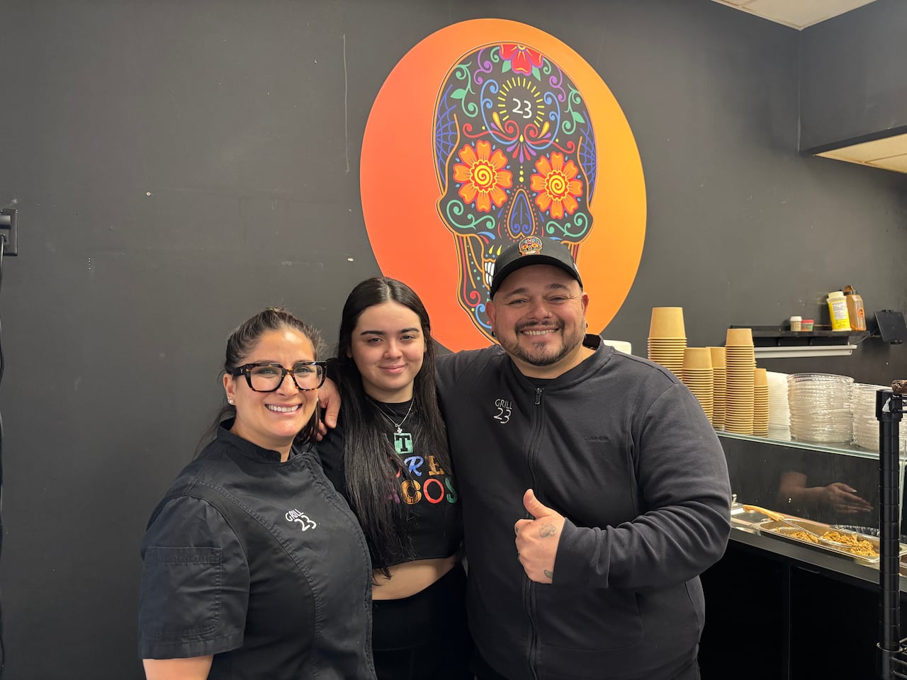 Gerry Rozo (on the right) and two women employees smile for a picture in front of the Grill 23 logo of a Calavera