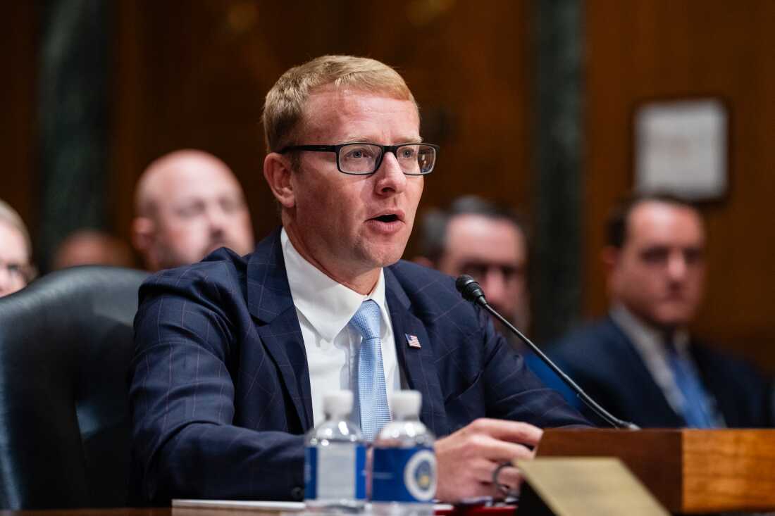 A man in a dark suit, white shirt and blue tie, with short hair and wearing glasses,  sits at a table in a congressional hearing room and speaks into a microphone. 