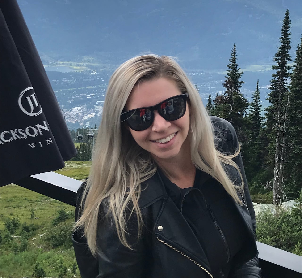 A white woman with long blond hair wearing a black jacket and black sunglasses stands in front of a mountain vista.