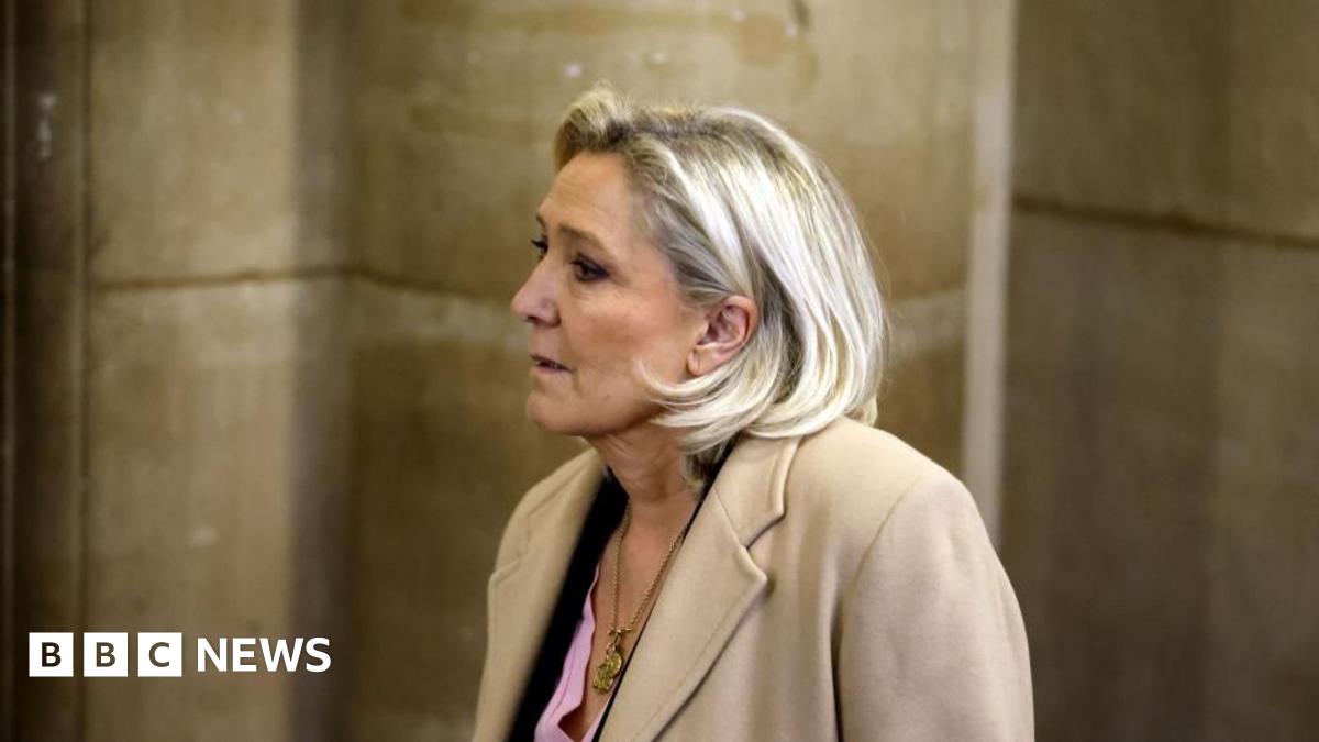 Millie Bobby Brown and David Harbour in character as Eleven and Jim Hopper. Eleven wears red shorts over grey leggings and a grey jumper. Jim Hopper wears a black leather jacket, a black beanie hat and dark trousers.They are speaking to director Frank Darabont on the set of Stranger Things. There is a vintage advertisement posted in the background with debrief on the ground.