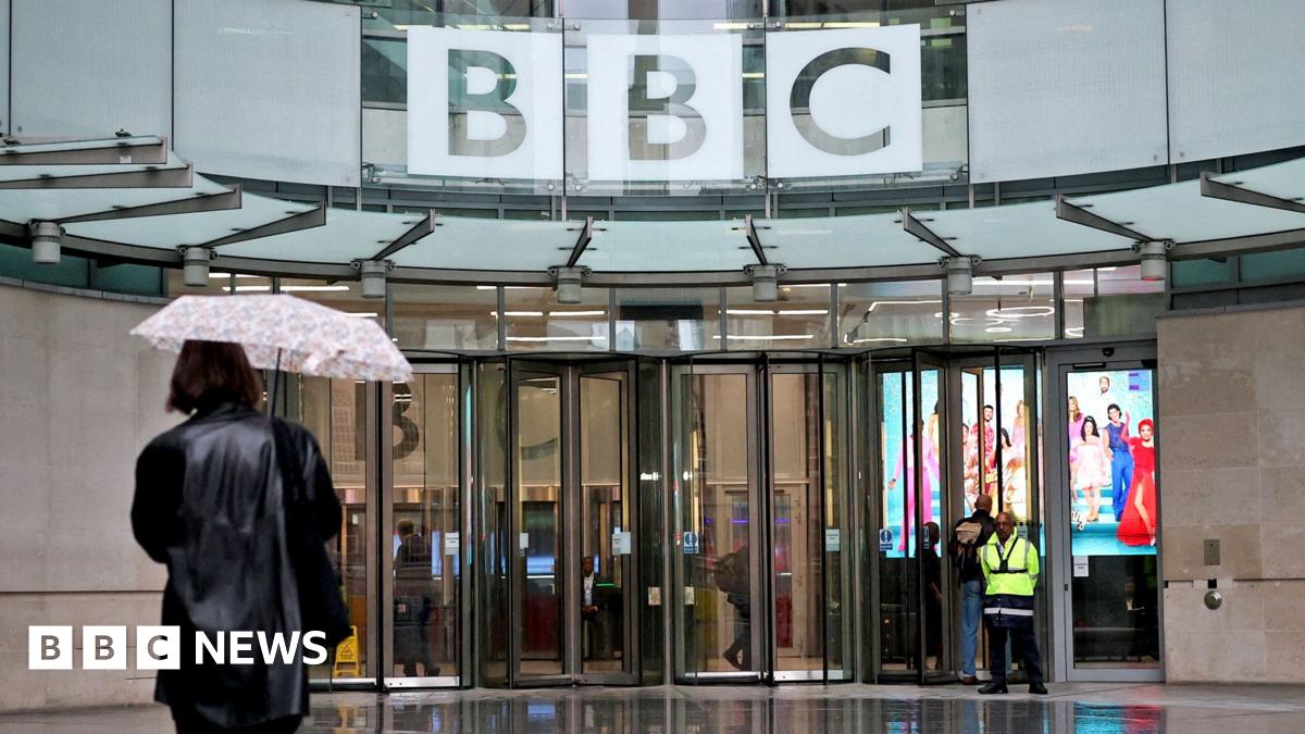 Millie Bobby Brown and David Harbour in character as Eleven and Jim Hopper. Eleven wears red shorts over grey leggings and a grey jumper. Jim Hopper wears a black leather jacket, a black beanie hat and dark trousers.They are speaking to director Frank Darabont on the set of Stranger Things. There is a vintage advertisement posted in the background with debrief on the ground.