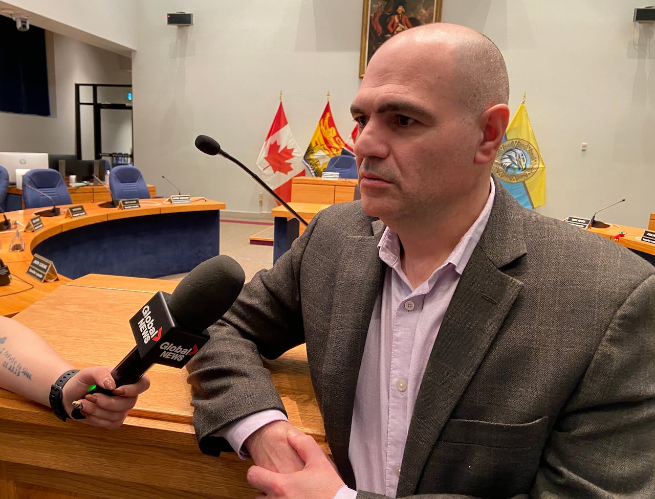 A man speaks into a microphone while standing in council chambers. 