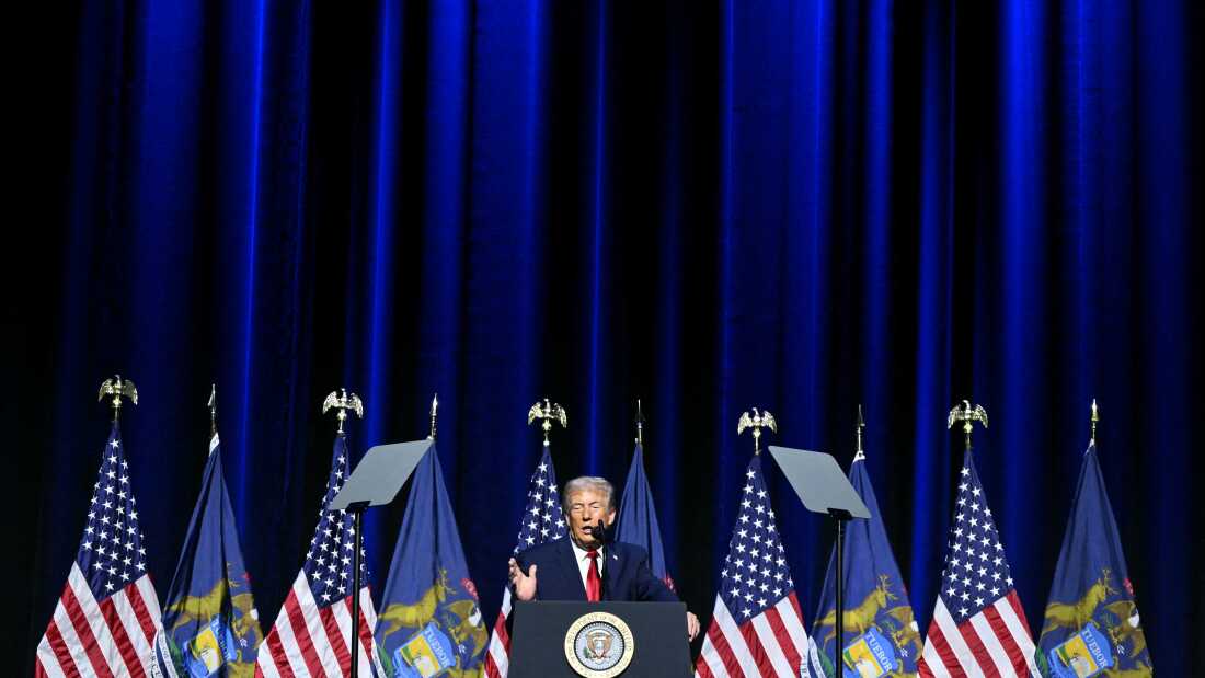 President Trump speaks at a podium at the Detroit Economic Club. He has several flags on either side of him with many of them being American flags. 