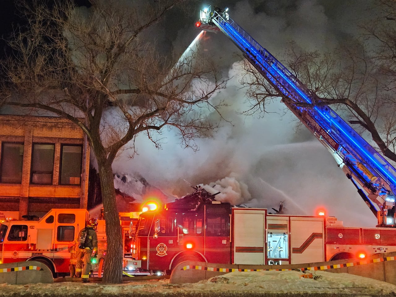 Thick smoke fills the air on a dark morning as fire crews in the foreground fight a building fire. An aerial ladder sprays water down.